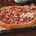 This photo shows a person's hand removing a slice from a whole Meat Lovers pizza.