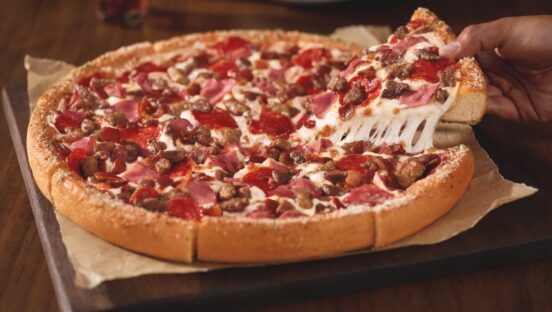 This photo shows a person's hand removing a slice from a whole Meat Lovers pizza.