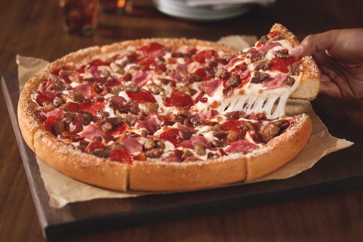 This photo shows a person's hand removing a slice from a whole Meat Lovers pizza.