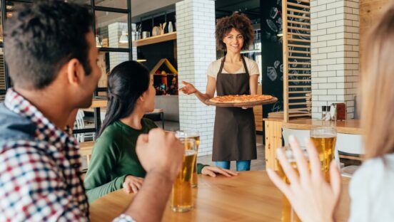 This photo shows an African american woman waitress bringing pizza to her customers.