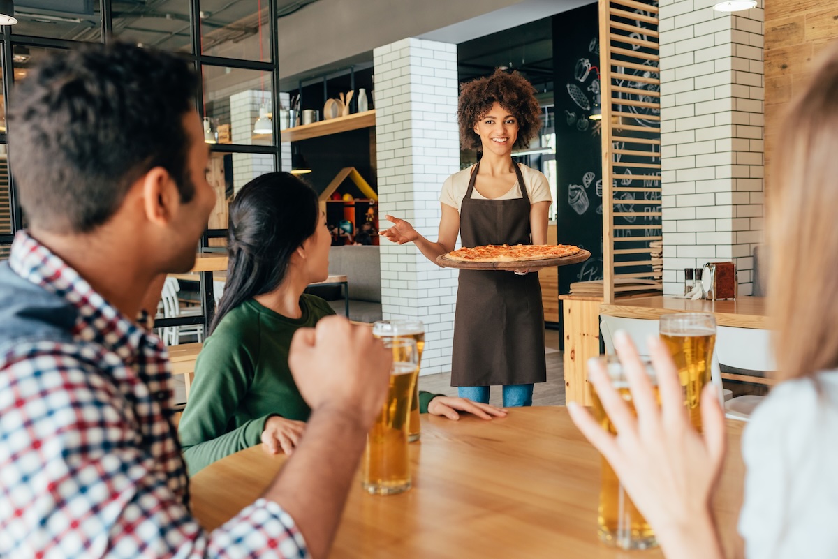 This photo shows an African american woman waitress bringing pizza to her customers.