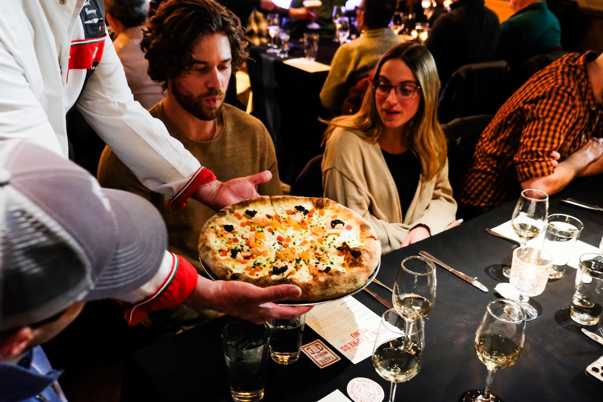 This photo shows Chef Robert Garvey presenting a pizza to a table of customers during the Orient Express ticketed dinner at Robert's Pizza and Dough Co.