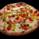 This photo shows a pizza topped with squash flowers, shrimp, tomatoes and mozzarella cheese.