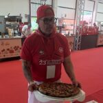 This photo shows Chris Battiste in red shirt and red backwards cap presenting a pizza to the judges.