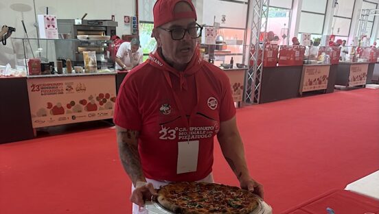 This photo shows Chris Battiste in red shirt and red backwards cap presenting a pizza to the judges.