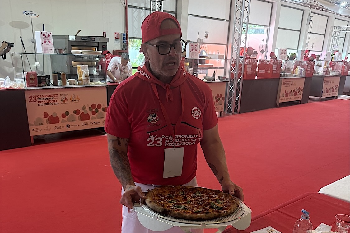 This photo shows Chris Battiste in red shirt and red backwards cap presenting a pizza to the judges.