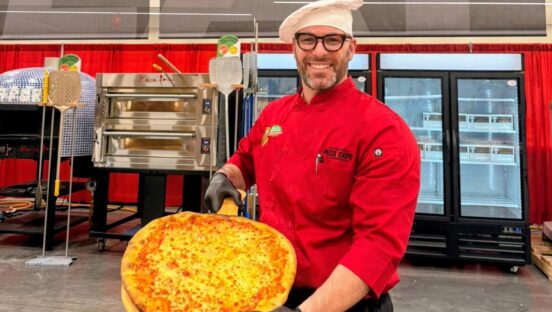 This photo shows Boris Berard wearing a red chef coat, a white chef hat and holding a large cheese pizza.