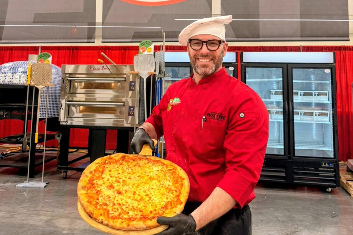 This photo shows Boris Berard wearing a red chef coat, a white chef hat and holding a large cheese pizza.