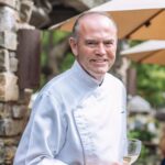 This photo shows Charlie Palmer in a white chef coat, smiling and holding a glass of white wine.