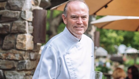 This photo shows Charlie Palmer in a white chef coat, smiling and holding a glass of white wine.