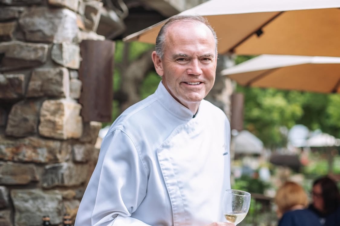 This photo shows Charlie Palmer in a white chef coat, smiling and holding a glass of white wine.