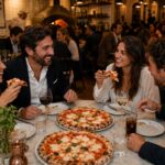 This photo shows a crowded pizzeria, with a table of two women and two men in the foreground eating Margherita pizzas.
