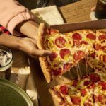 This photo shows a woman's hands pulling a slice from a whole New York-style pizza.