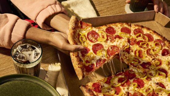 This photo shows a woman's hands pulling a slice from a whole New York-style pizza.