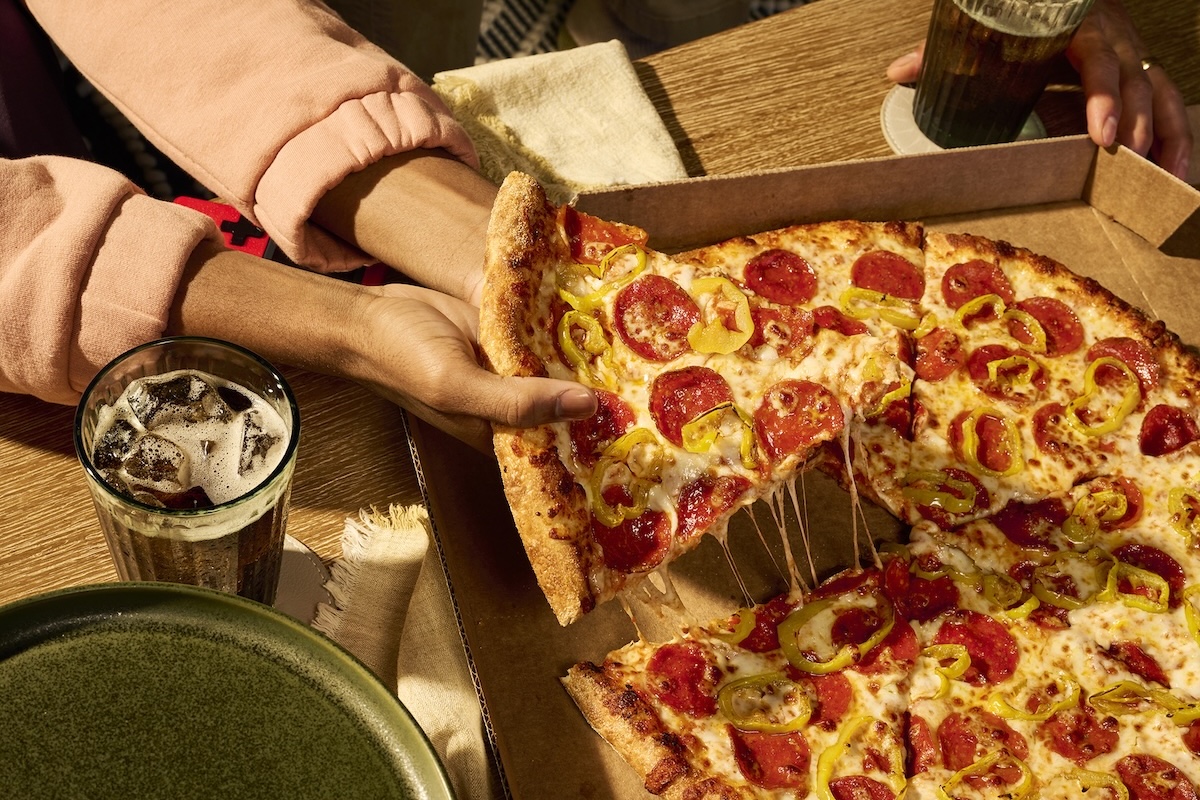 This photo shows a woman's hands pulling a slice from a whole New York-style pizza.