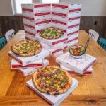 This photo shows three different pizzas arranged at different levels on a table, and behind it is a stack of branded pizza boxes.