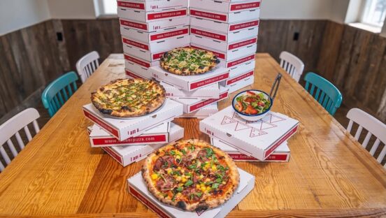 This photo shows three different pizzas arranged at different levels on a table, and behind it is a stack of branded pizza boxes.