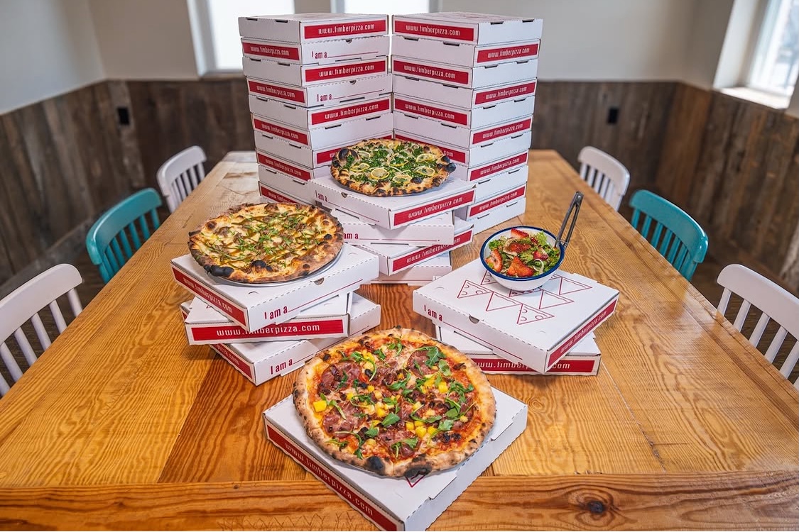 This photo shows three different pizzas arranged at different levels on a table, and behind it is a stack of branded pizza boxes.