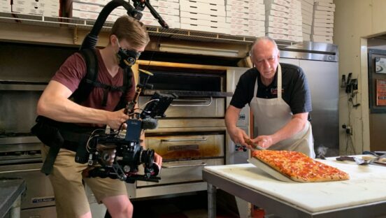 This photo shows an influencer with extensive camera gear standing near a prep table as a pizza maker gets ready to put a pizza in the oven.
