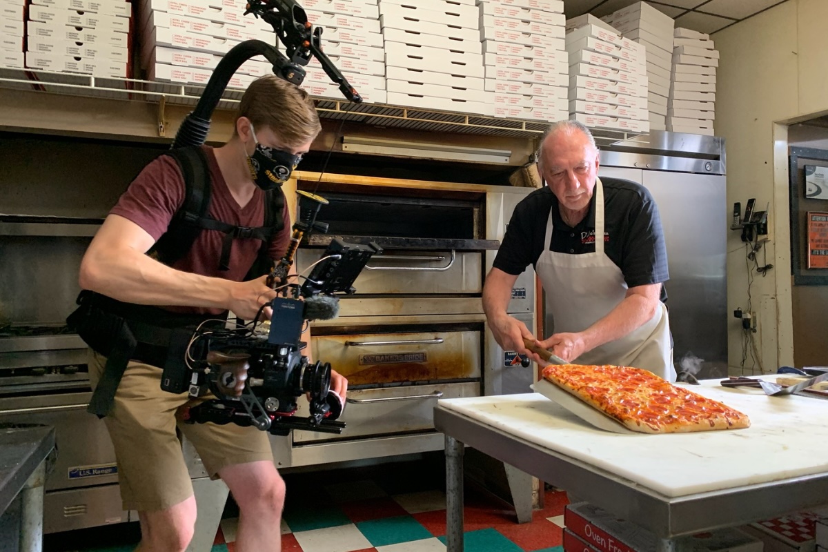 This photo shows an influencer with extensive camera gear standing near a prep table as a pizza maker gets ready to put a pizza in the oven.