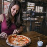 This photo shows a woman in a red tank top smiling and getting ready to pull a slice from a whole Margherita pizza.