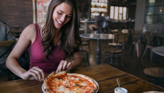 This photo shows a woman in a red tank top smiling and getting ready to pull a slice from a whole Margherita pizza.