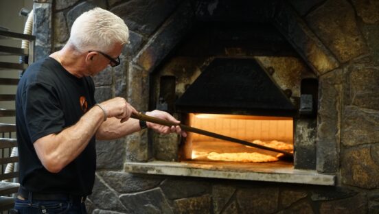 This photo shows Bill Pustari moving a pizza around his coal-fired oven with a peel.
