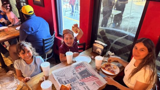 This photo shows a woman and two little girls at Big Apple Pizza. The two children are all smiles, and the girl on the right is excitedly holding up a slice of pizza.