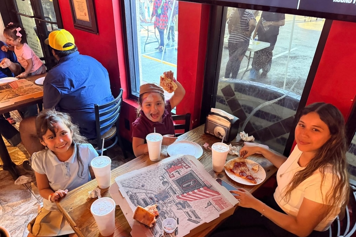 This photo shows a woman and two little girls at Big Apple Pizza. The two children are all smiles, and the girl on the right is excitedly holding up a slice of pizza.