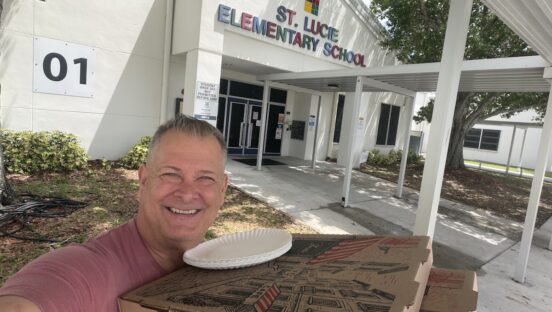 This photo, a selfie, shows Scott VanDuzer smiling and holding a bunch of pizza boxes as he stands in front of St. Lucie Elementary School.
