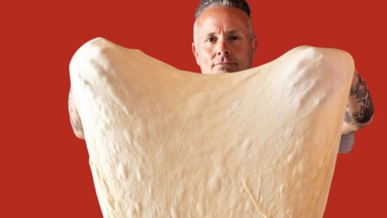This photo shows Tony Gemignani stretching a gigantic piece of dough into the shape of a heart.