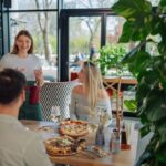 This photo shows a cheerful young waitress explaining menu to customers sitting at a table, ready to eat pizza in a pizzeria restaurant