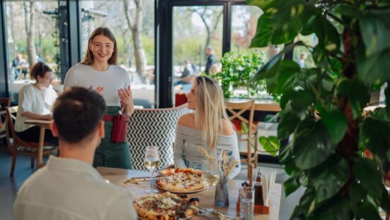 This photo shows a cheerful young waitress explaining menu to customers sitting at a table, ready to eat pizza in a pizzeria restaurant