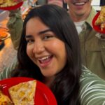 This photo shows a young woman holding a plate with two pizza slices up to the camera and smiling happily.