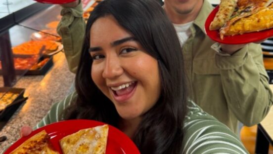 This photo shows a young woman holding a plate with two pizza slices up to the camera and smiling happily.
