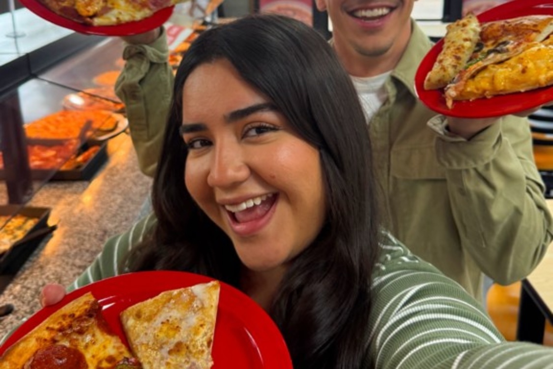 This photo shows a young woman holding a plate with two pizza slices up to the camera and smiling happily.