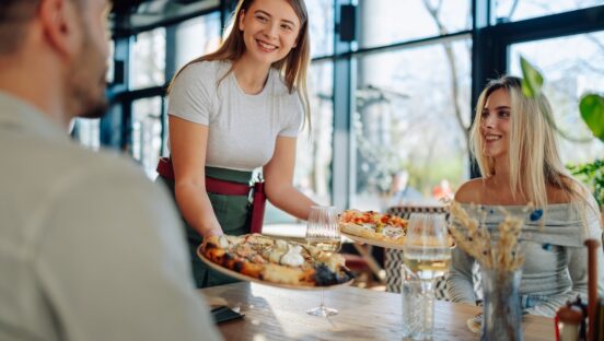 This photo shows a Smiling waitress bringing freshly baked pizzas to young customers at a table in a modern pizzeria