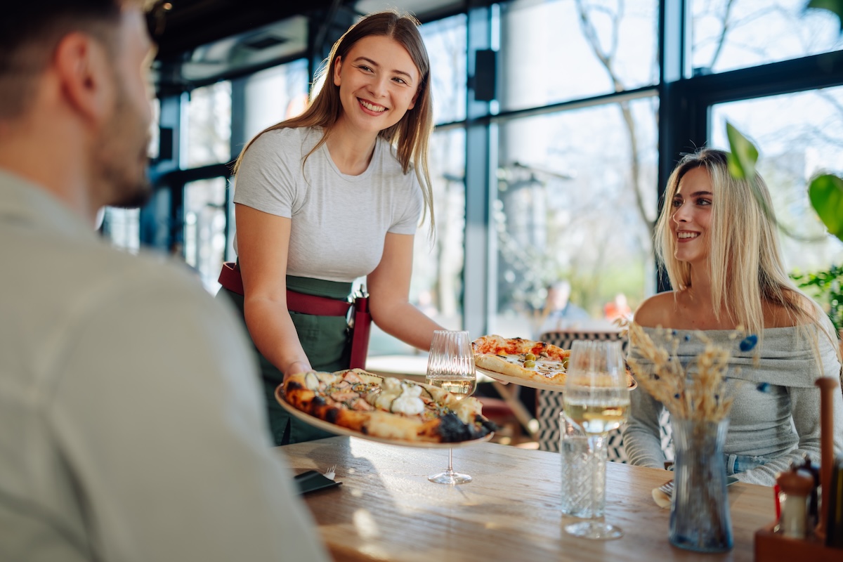 This photo shows a Smiling waitress bringing freshly baked pizzas to young customers at a table in a modern pizzeria