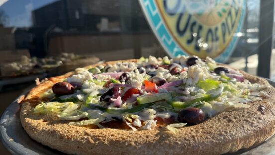 This photo shows a a thicker-crust pizza with lettuce, tomatoes, Kalamata olives, red onions, green peppers and crumbled feta cheese.