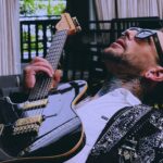 This photo shows Navarone Garcia, wearing sunglasses and a stylish jacket and shirt, leaning back and holding a guitar while gazing upwards through sunglasses.