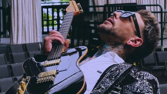 This photo shows Navarone Garcia, wearing sunglasses and a stylish jacket and shirt, leaning back and holding a guitar while gazing upwards through sunglasses.