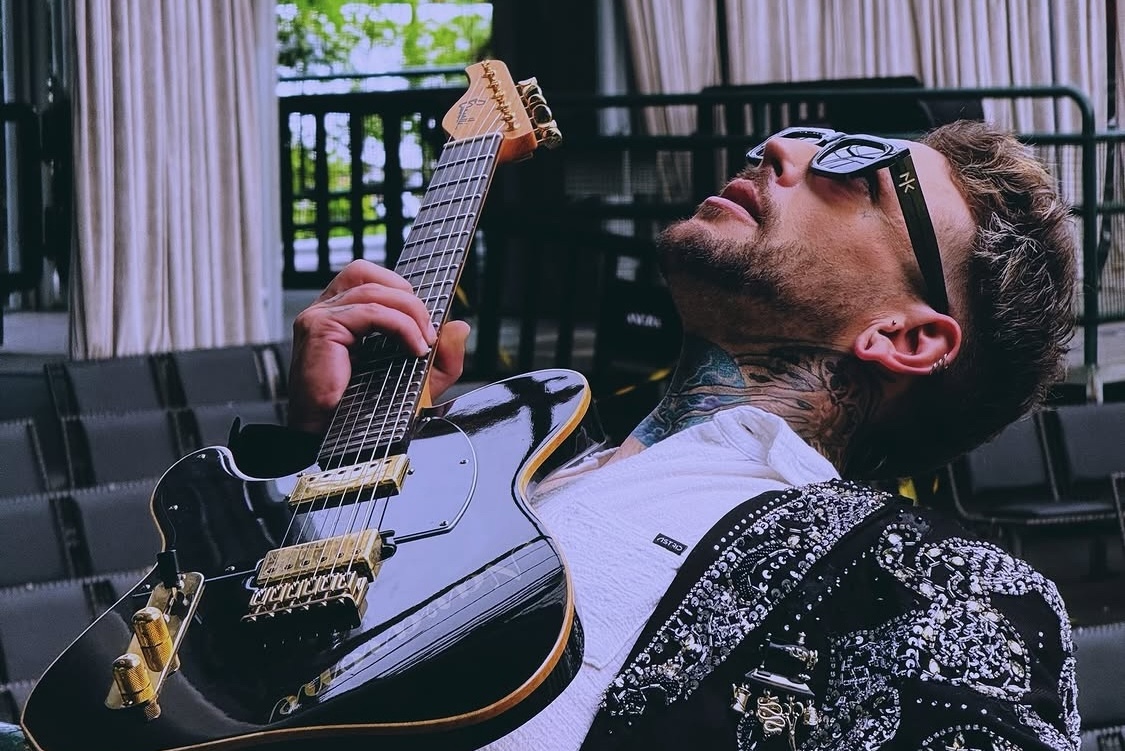 This photo shows Navarone Garcia, wearing sunglasses and a stylish jacket and shirt, leaning back and holding a guitar while gazing upwards through sunglasses.