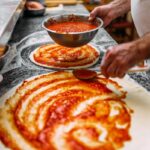 Chef baker making pizza at the kitchen. Adding tomato sauce on a pizza dough