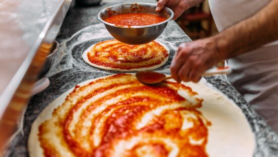 Chef baker making pizza at the kitchen. Adding tomato sauce on a pizza dough