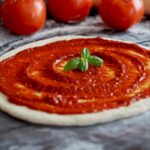 This photo shows an unbaked pizza crust with tomato sauce and three fresh whole tomatoes set off to the side.