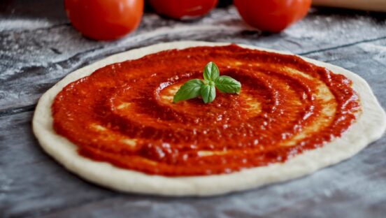 This photo shows an unbaked pizza crust with tomato sauce and three fresh whole tomatoes set off to the side.