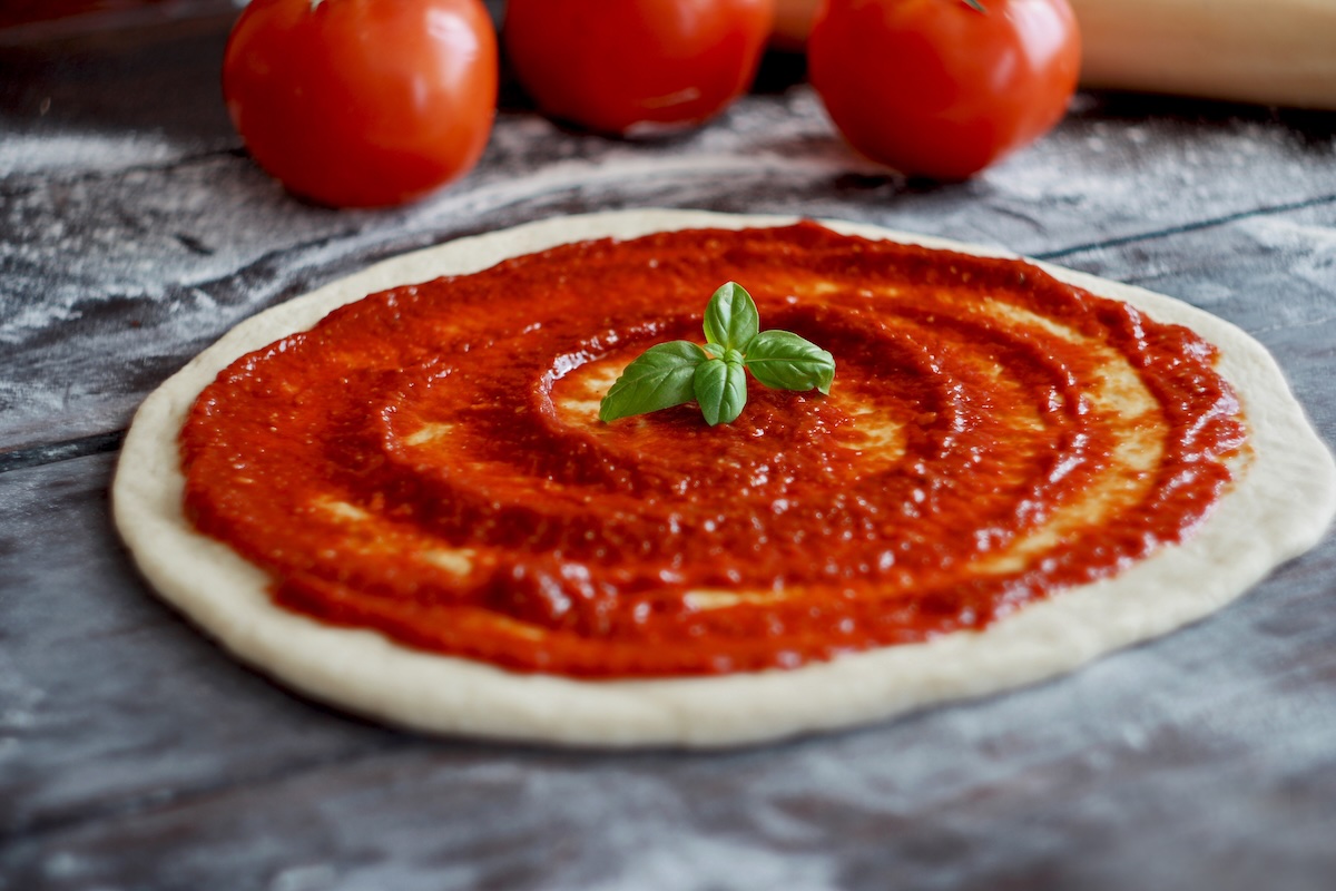 This photo shows an unbaked pizza crust with tomato sauce and three fresh whole tomatoes set off to the side.