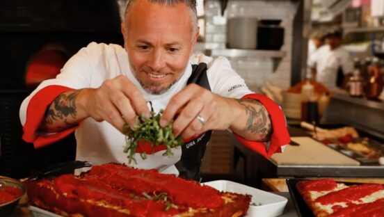 This photo shows Tony Gemignani leaning in closely and adding arugula to a Detroit-style pizza.