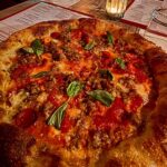 This photo shows a round pizza with a thick cornicione topped with sausage and pepperoni.