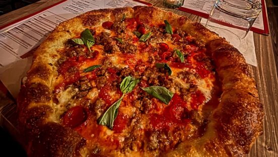 This photo shows a round pizza with a thick cornicione topped with sausage and pepperoni.
